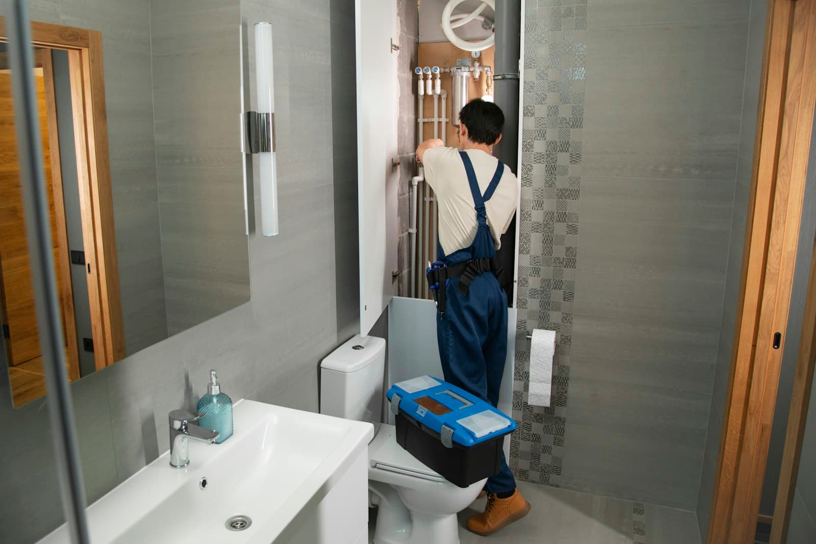 Plumber in blue overalls servicing wall-mounted bathroom pipework